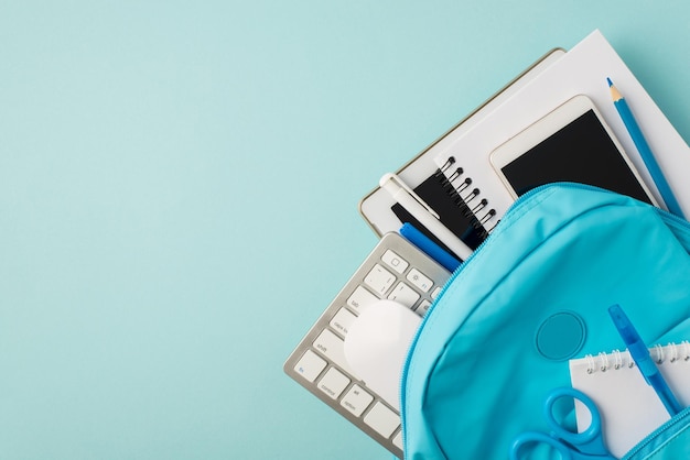 Bovenaanzicht foto van tas met tablet computer smartphone notitieblokken potlood pennen toetsenbord muis en schaar op geïsoleerde pastel blauwe achtergrond met copyspace