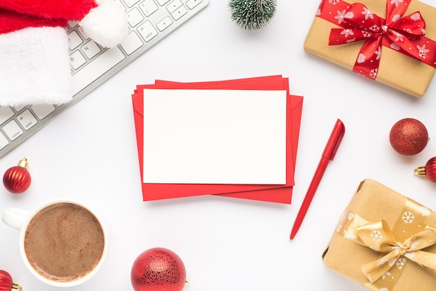 Bovenaanzicht foto van toetsenbord kopje warm drinken kerstman hoed grenen speelgoed rode kerstboom ballen geschenkdozen stapel rode enveloppen papier blad en pen op geïsoleerde witte achtergrond met copyspace