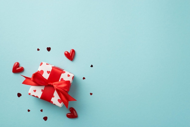 Bovenaanzicht foto van Valentijnsdag decor geschenkdoos in wit inpakpapier met hart patroon en rode strik omringd door kleine hartjes en confetti op geïsoleerde pastel blauwe achtergrond met copyspace