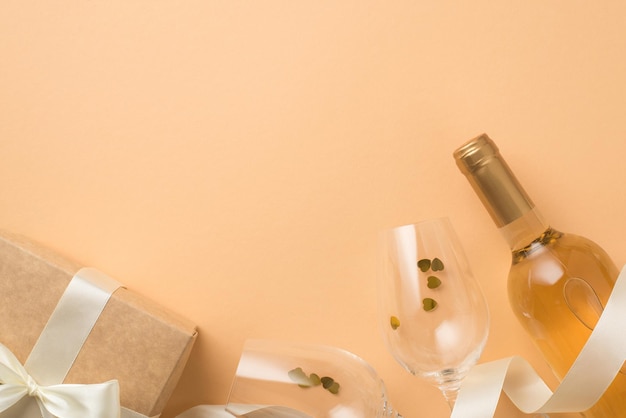 Bovenaanzicht foto van valentijnsdag decoraties wijnfles wijnglazen met hartvormige confetti satijnen lint ambachtelijke papier geschenkdoos met witte strik op geïsoleerde pastel beige achtergrond met copyspace