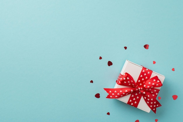 Bovenaanzicht foto van Valentijnsdag decoraties witte geschenkdoos met rode strik en hartvormige confetti op geïsoleerde pastelblauwe achtergrond met lege ruimte