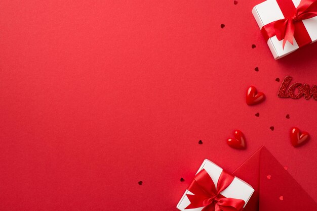 Bovenaanzicht foto van Valentijnsdag decoraties witte geschenkdozen met rode strikken envelop kleine harten inscriptie liefde en confetti op geïsoleerde rode achtergrond met lege ruimte