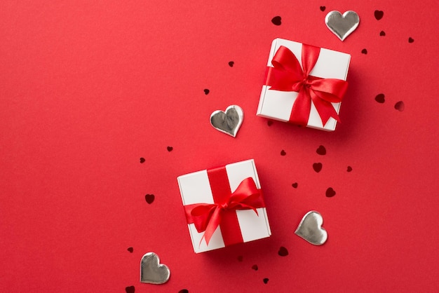 Bovenaanzicht foto van Valentijnsdag decoraties witte geschenkdozen met rood lint strikken decoratieve zilveren harten en confetti op geïsoleerde rode achtergrond met lege ruimte