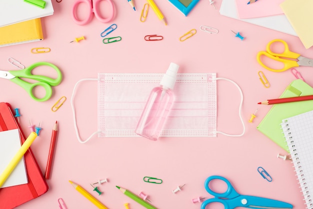 Bovenaanzicht foto van veelkleurige briefpapier schaar pennen potloden pushpins clips rekenmachine schriften en ontsmettingsmiddel fles op medisch gezichtsmasker in het midden geïsoleerde pastel roze achtergrond