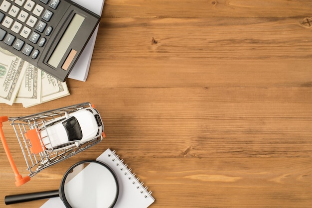 Bovenaanzicht foto van werkplek met papieren organisator rekenmachine vergrootglas dollars en automodel in winkelwagen model op geïsoleerde houten tafel achtergrond met copyspace