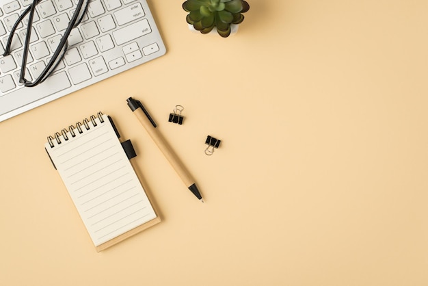 Bovenaanzicht foto van witte toetsenbord bril plant schrijfboek pen en bindmiddelen op geïsoleerde beige achtergrond met copyspace