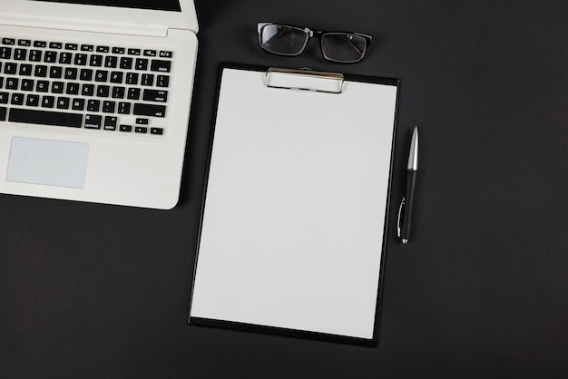 Bovenaanzicht foto van zwarte klembord map met wit papier blad pen bril en laptop op geïsoleerde zwarte achtergrond met lege ruimte