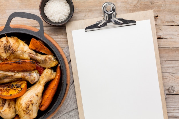 Bovenaanzicht gebakken kip en groenten in pan met leeg klembord