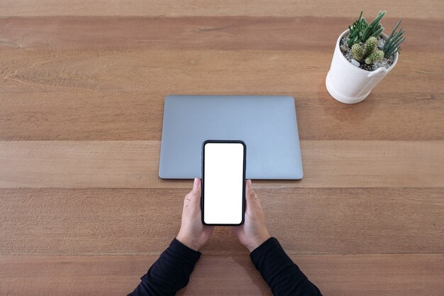 Bovenaanzicht mockup afbeelding van een handen met een leeg wit scherm mobiele telefoon en laptop op houten tafel in kantoor