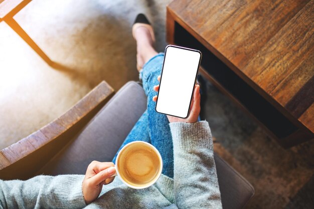 Bovenaanzicht mockup afbeelding van een vrouw die een mobiele telefoon vasthoudt met een leeg wit desktopscherm terwijl ze koffie drinkt