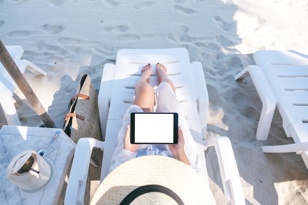 Bovenaanzicht mockup afbeelding van een vrouw die een zwarte tablet-pc vasthoudt en gebruikt met een leeg bureaubladscherm terwijl ze op een strandstoel op het strand ligt