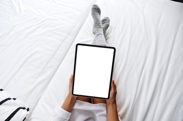 Bovenaanzicht mockup-afbeelding van een vrouw die een zwarte tablet-pc vasthoudt met een leeg wit bureaubladscherm terwijl ze thuis op een gezellig wit bed ligt