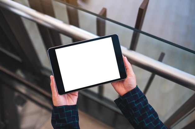 Bovenaanzicht mockup-afbeelding van een vrouw die een zwarte tablet-pc vasthoudt met een leeg wit desktopscherm binnenshuis