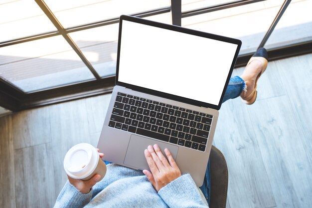 Bovenaanzicht mockup-afbeelding van een vrouw die het touchpad van een laptop gebruikt en aanraakt met een leeg wit bureaubladscherm terwijl ze koffie drinkt