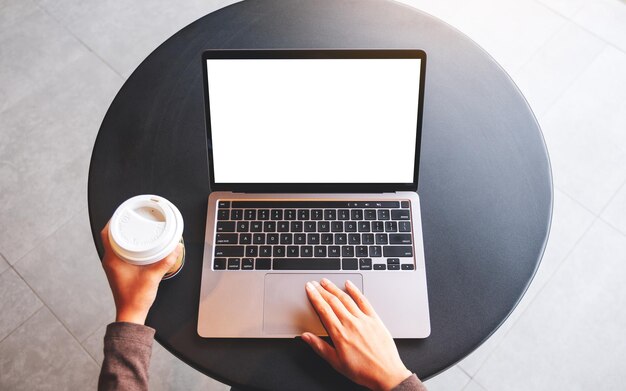 Bovenaanzicht mockup afbeelding van een vrouw die het touchpad van een laptop gebruikt en aanraakt met een leeg wit desktopscherm terwijl ze koffie drinkt