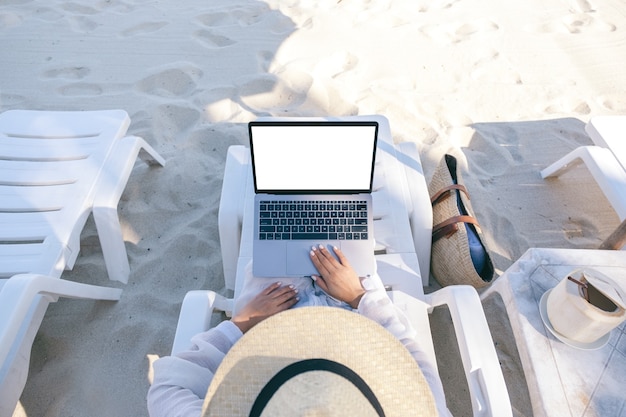 Bovenaanzicht mockup afbeelding van een vrouw die laptopcomputer gebruikt en typt met een leeg bureaublad terwijl ze op de strandstoel op het strand ligt