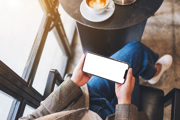 Bovenaanzicht mockup afbeelding van een vrouw die zwarte mobiele telefoon vasthoudt met een leeg desktopscherm terwijl ze in café zit