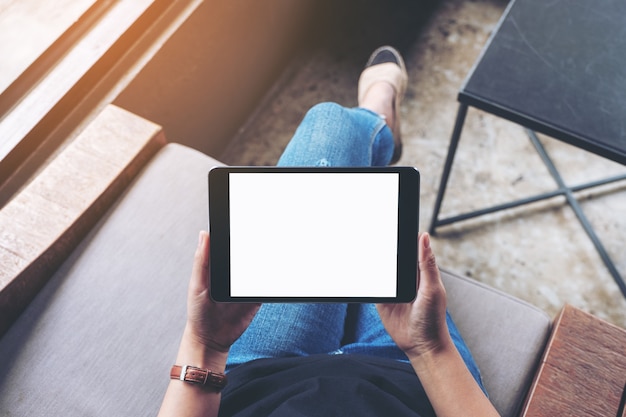 Bovenaanzicht mockup afbeelding van een vrouw die zwarte tablet-pc vasthoudt met een leeg wit scherm terwijl ze in café zit