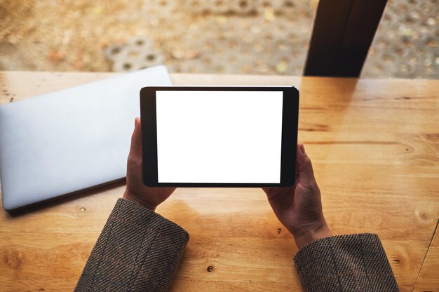 Bovenaanzicht mockup afbeelding van een vrouw die zwarte tablet-pc zit en vasthoudt met een leeg wit desktopscherm met laptop op tafel