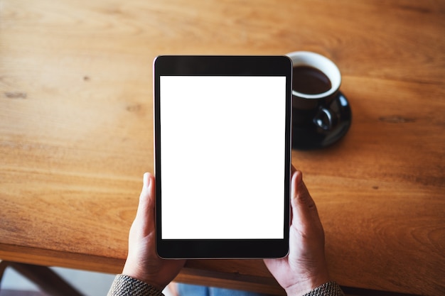 Bovenaanzicht mockup afbeelding van handen met zwarte tablet pc met leeg wit scherm met koffiekopje op houten tafel