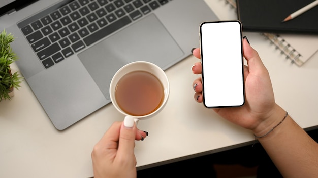 Bovenaanzicht Moderne kantoortafel met vrouwenhanden met theekopje en smartphone leeg schermmodel