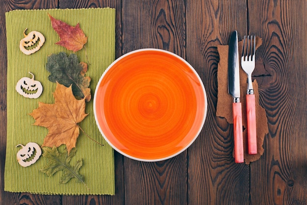 Bovenaanzicht op een Halloween-achtergrond met een lege oranje plaat klaar voor het plaatsen van gekleurde bladeren van voedselbestek op een houten tafel en spookachtige, uit hout gesneden pompoenen kopiëren ruimte