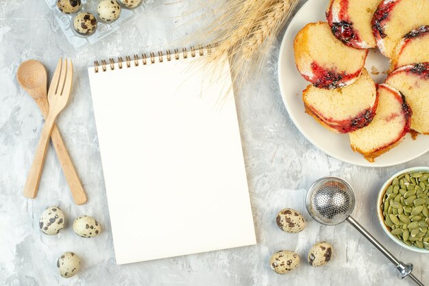 Bovenaanzicht plakjes jam cake op bord andere dingen in kommen tarwe spikes notitieblok houten vork en lepel op tafel