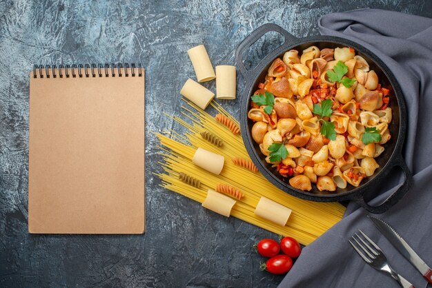 bovenaanzicht smakelijke pasta in koekenpan spaghetti rigatoni spiralen kerstomaatjes mes en vork notitieboekje op grijze achtergrond