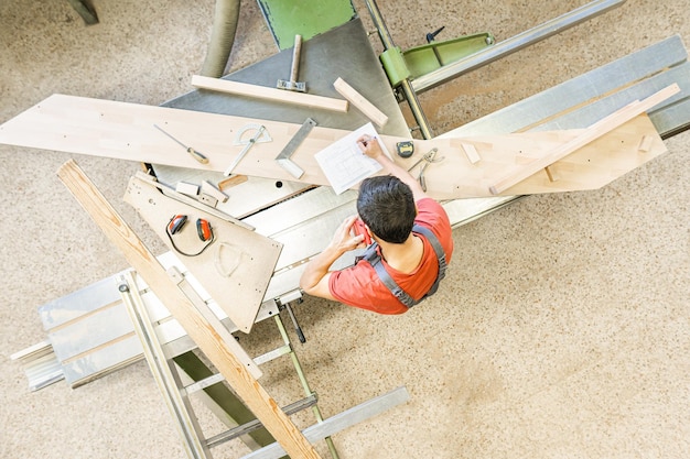 Bovenaanzicht van anonieme mannelijke timmerman die een telefoongesprek voert terwijl hij aantekeningen maakt bij een schuiftafelzaag in professioneel licht schrijnwerk