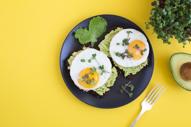 Bovenaanzicht van avocadotoosts voor het ontbijt met roggebrood, avocadopuree, gebakken eieren en microgroen op de gele achtergrond. Ruimte kopiëren.