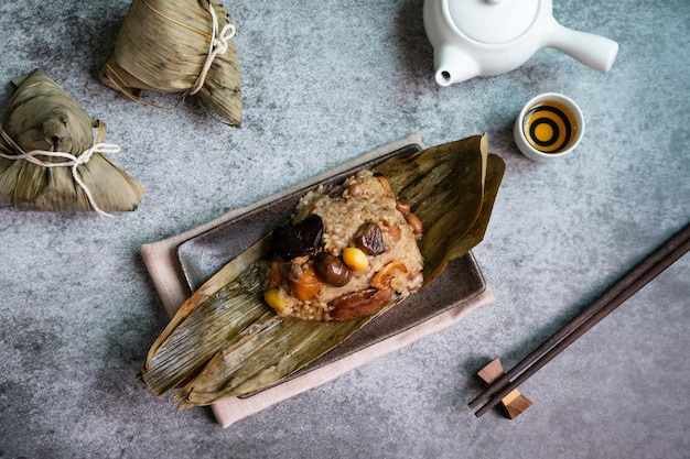 Bovenaanzicht van aziatische smakelijke zelfgemaakte gerechten in drakenboot (duan wu) festival, rijst knoedels of zongzi verpakt door gedroogde bamboebladeren op plaat met thee op zwarte bord