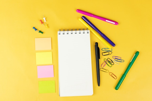 Bovenaanzicht van blanco notebook en schoolbenodigdheden zoals gekleurde markeringen, sticker en clipers op gele achtergrond, ruimte voor tekst.