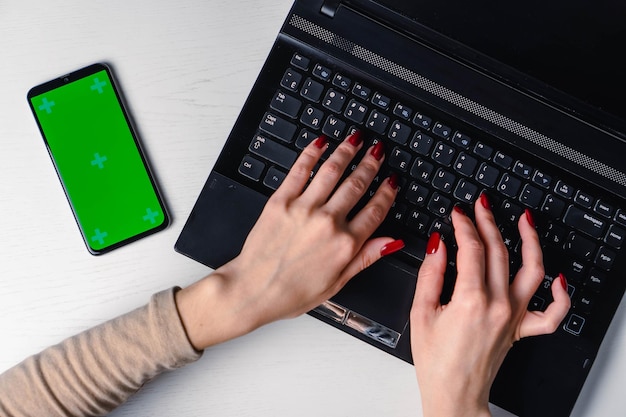 Bovenaanzicht van dames handen typen op laptop toetsenbord geplaatst op witte desktop met lege smartphon
