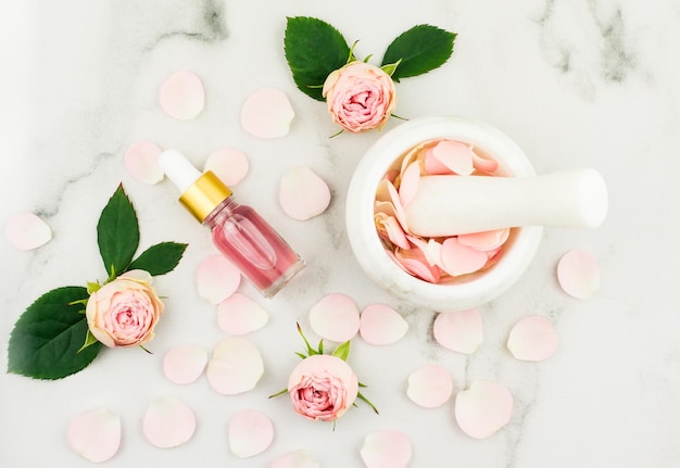 Bovenaanzicht van de fles met roze cosmetische olie, vijzel en stamper met rozenblaadjes, bloemen, groene bladeren.