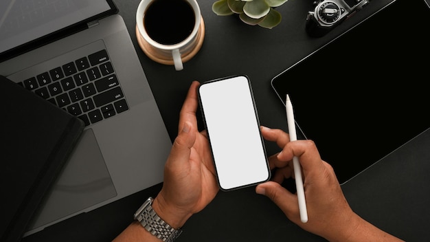 Bovenaanzicht van de handen van een man met een mockup met een wit scherm van een smartphone op een moderne donkere werktafel
