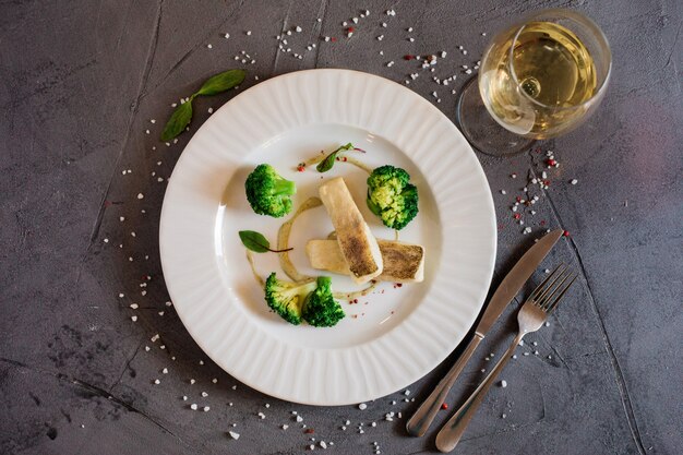 Bovenaanzicht van de heerlijke lunch met witte vis en broccoli op het bord en een glas wijn of sap.