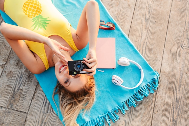 Bovenaanzicht van een aantrekkelijk lachend jong blond meisje in zwembroek ontspannen op een zomerdeken op het strand over houten vloer, met fotocamera
