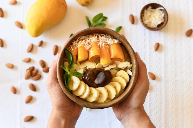 Bovenaanzicht van een anonieme persoon die een houten kom vasthoudt met lekkere plakjes banaan en mango, zaden en muntblaadjes tegen een wit tafelkleed