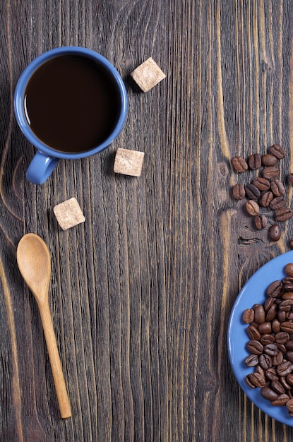 Bovenaanzicht van een blauwe kop koffie, schotel met bonen, suikerklontjes en een lepel op een donkere houten achtergrond met ruimte voor tekst