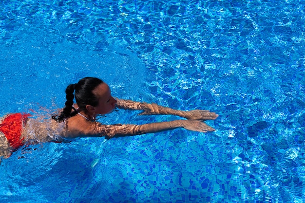 Bovenaanzicht van een gebruind meisje, vrouw, model in een rode zwembroek, zwemmen in het blauwe water van het zwembad.