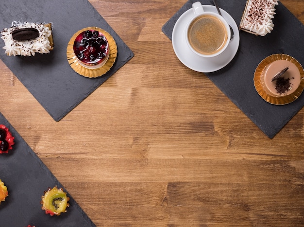 Bovenaanzicht van een kopje koffie met verschillende taarten met verschillende smaken over een houten tafel. Taarten met puddingroom. Taart met witte room en lekker koekje bovenop.