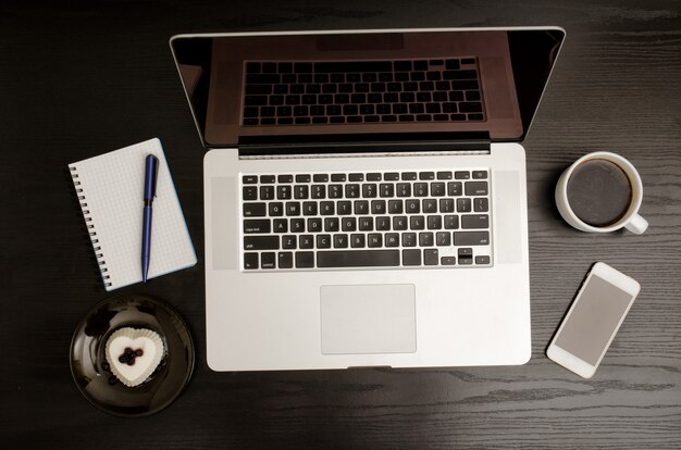 Bovenaanzicht van een laptop toetsenbord, koffiemok, een smartphone, een laptop en een dessert op een zwarte houten tafel