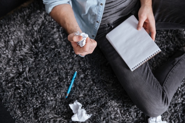 Bovenaanzicht van een man die verfrommeld papier en notitieblok vasthoudt terwijl hij op tapijt zit