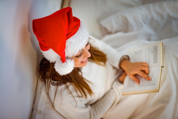 Bovenaanzicht van een mooi lachend meisje met kerstmuts ontspannen in een gezellig bed en leesboek.