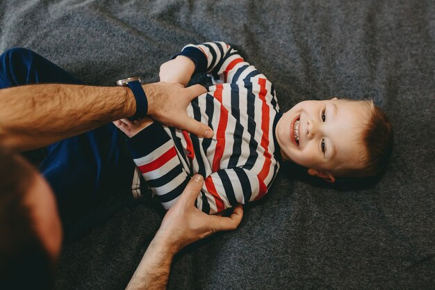 Bovenaanzicht van een mooie gelukkige jongen, leunend op een bed, lachen tijdens het spelen met zijn vader in de ochtend.