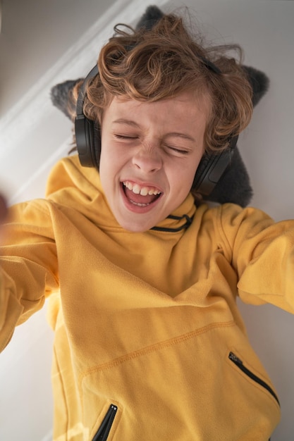 Bovenaanzicht van een opgetogen preteenjongen in een hoofdtelefoon die met gesloten ogen zingt terwijl hij naar muziek luistert en een zelfportret maakt