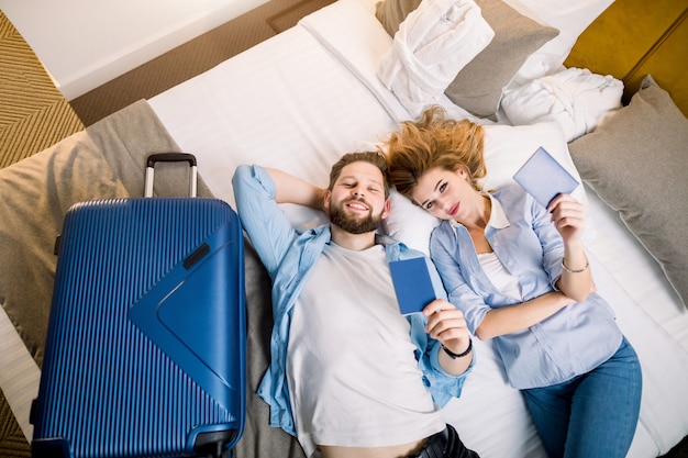 Bovenaanzicht van een paar toeristen, mooie vrouw en knappe man in vrijetijdskleding, liggend op bed in hotelkamer, met hun paspoorten. Reizen, hotel, boekingsconcept