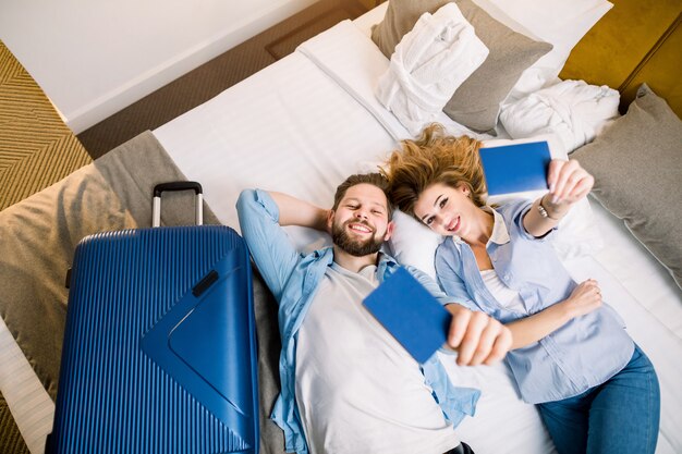 Bovenaanzicht van een paar toeristen, mooie vrouw en knappe man in vrijetijdskleding, liggend op bed in hotelkamer, met hun paspoorten. Reizen, hotel, boekingsconcept