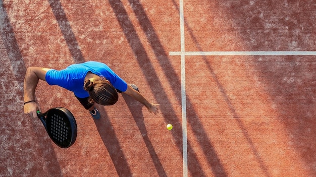 Bovenaanzicht van een paddle-tennisser die de bal stuitert voor een service op een buitenbaan.