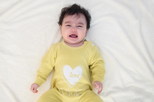 Bovenaanzicht van een schattig klein aziatisch babymeisje met een gele jurk, liggend op het bed, verdrietig en huilend